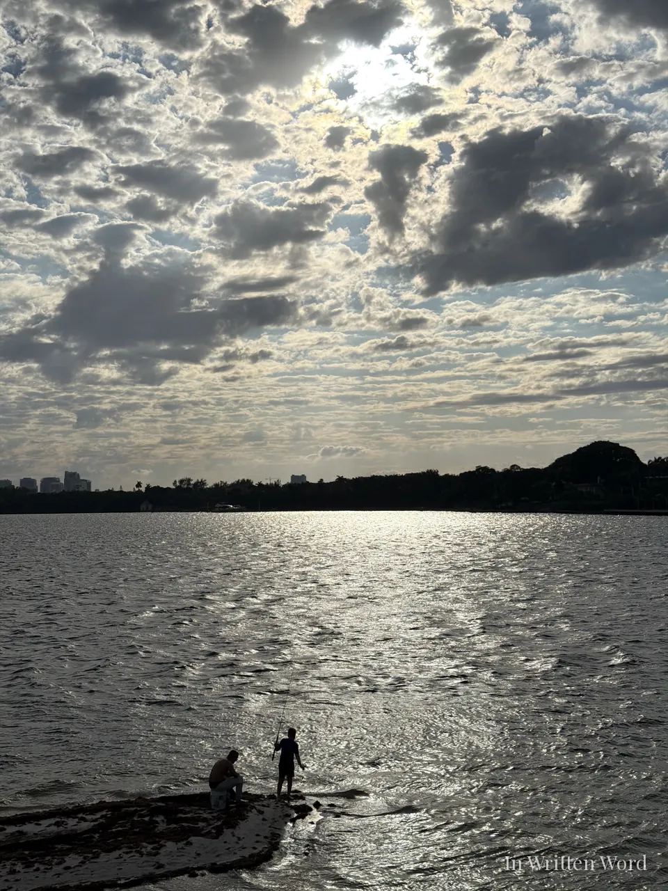 Two figures standing on a narrow shoreline looking out over shimmering water under a cloud-filled sky in Miami
