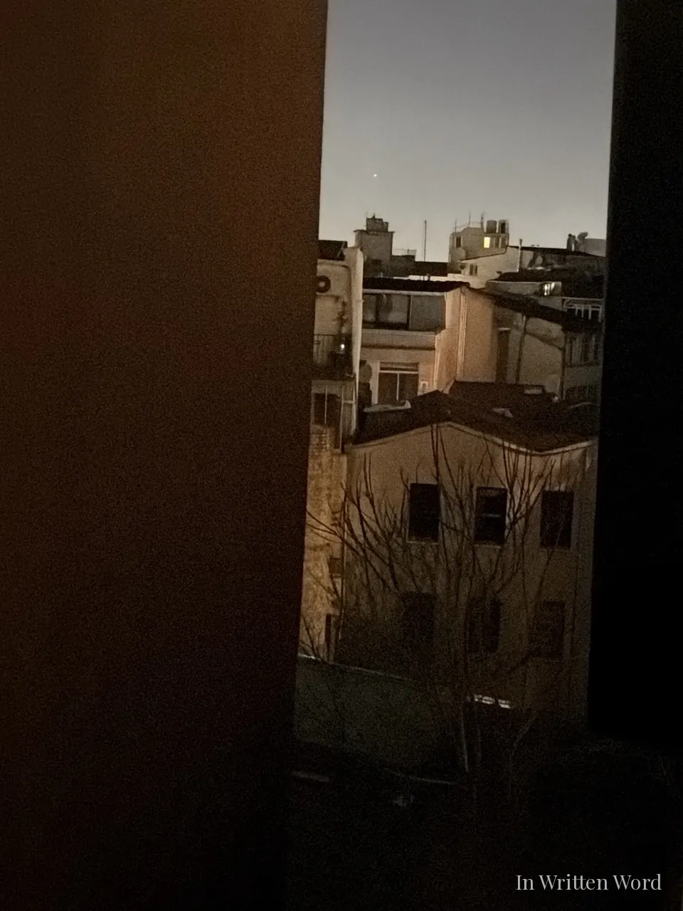  A narrow view through a hotel room window in Istanbul at night, showing rooftops, a bare winter tree, and a single star in the blue-grey sky