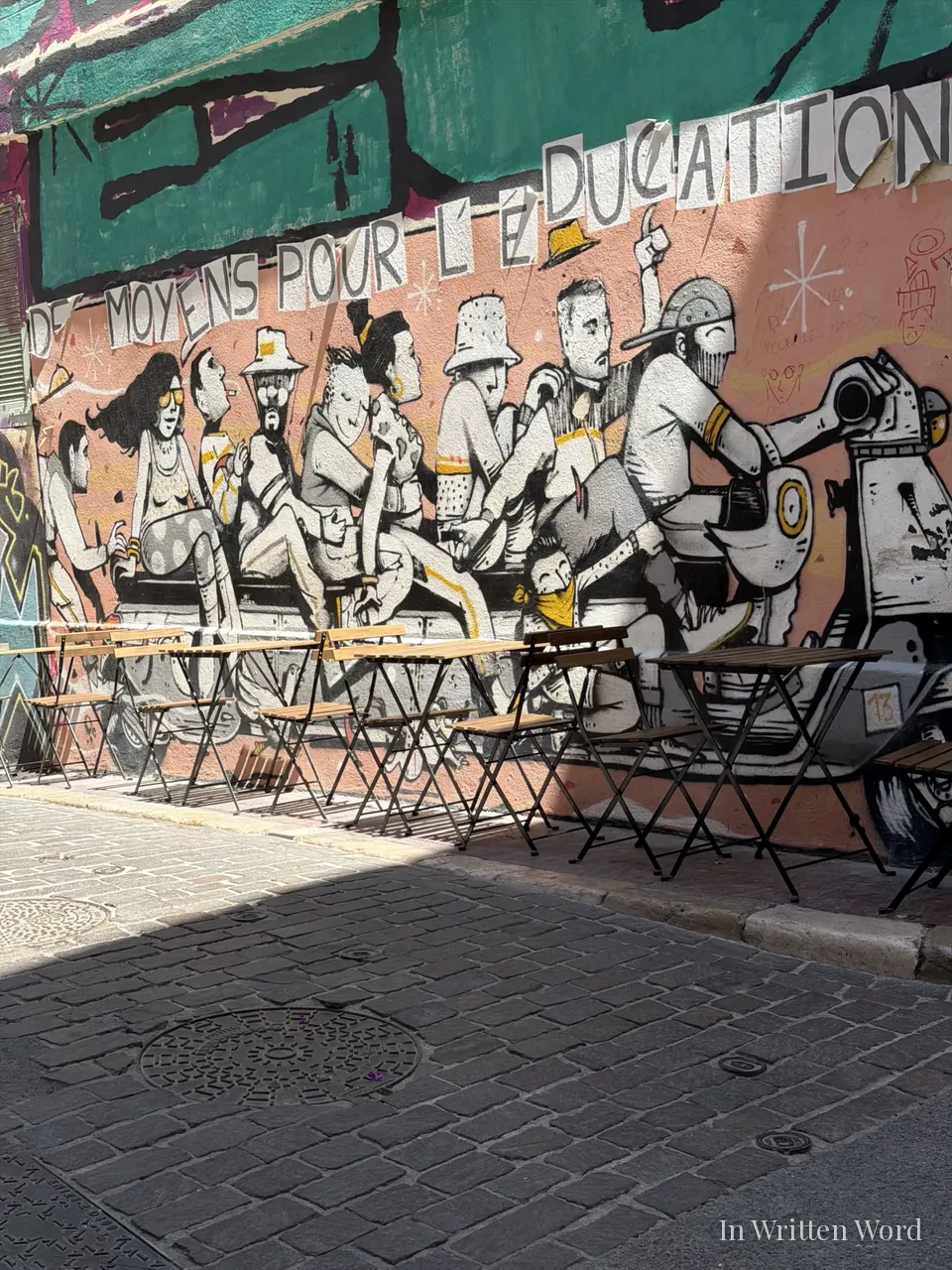 A large street mural of figures crowded onto a scooter, with café tables and folding chairs lined up beneath it on a cobblestone street in Marseille.