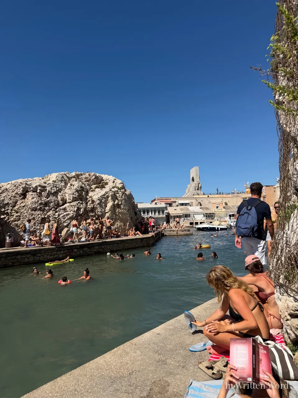 People swimming in calm green water below a large limestone rock along Marseille’s coastline, with sunbathers lining the stone ledge.