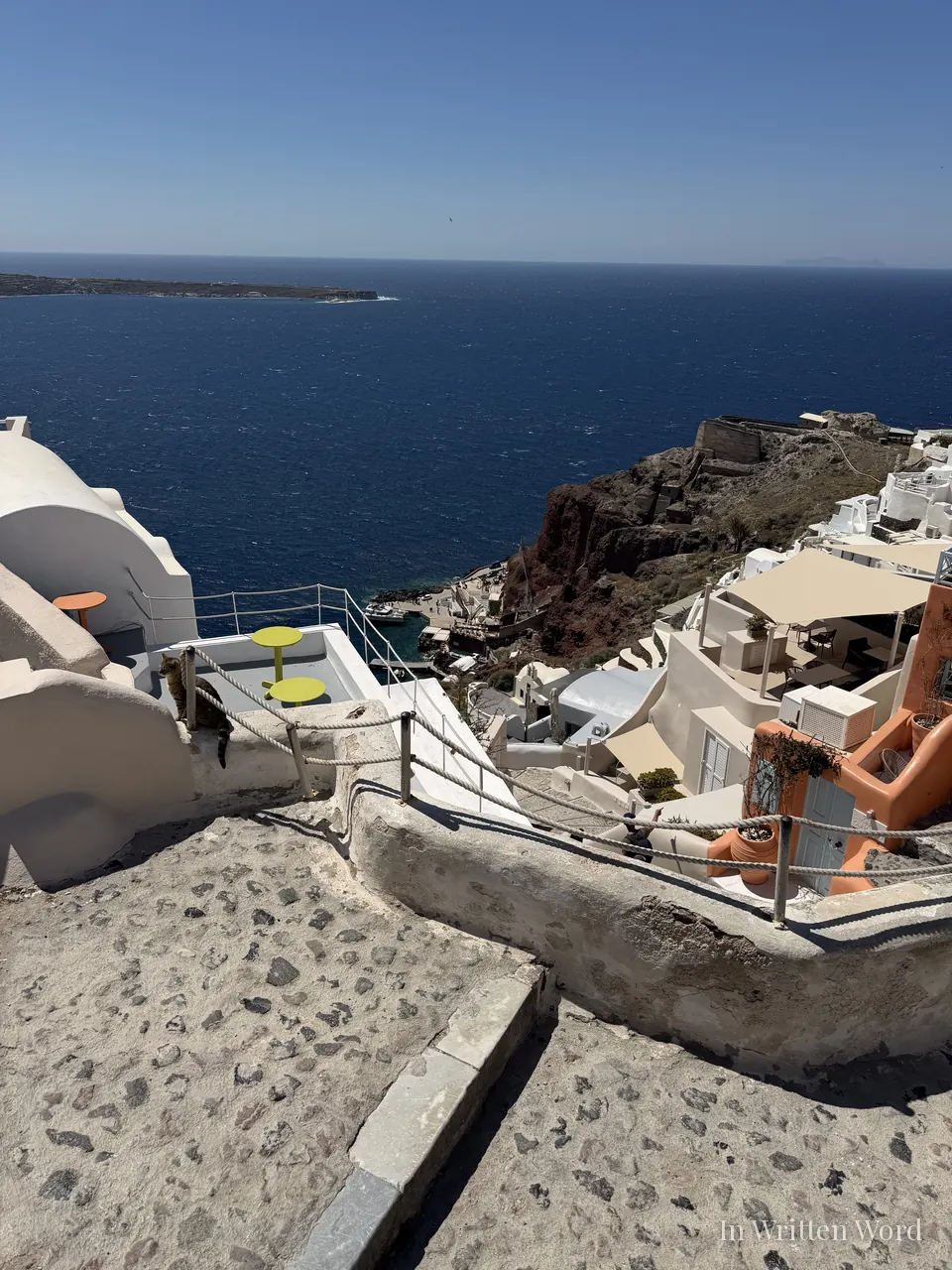 View of the Santorini caldera from a cliffside terrace, with a tabby cat resting on a low white wall, yellow café tables, and whitewashed buildings descending toward the deep blue Aegean.
