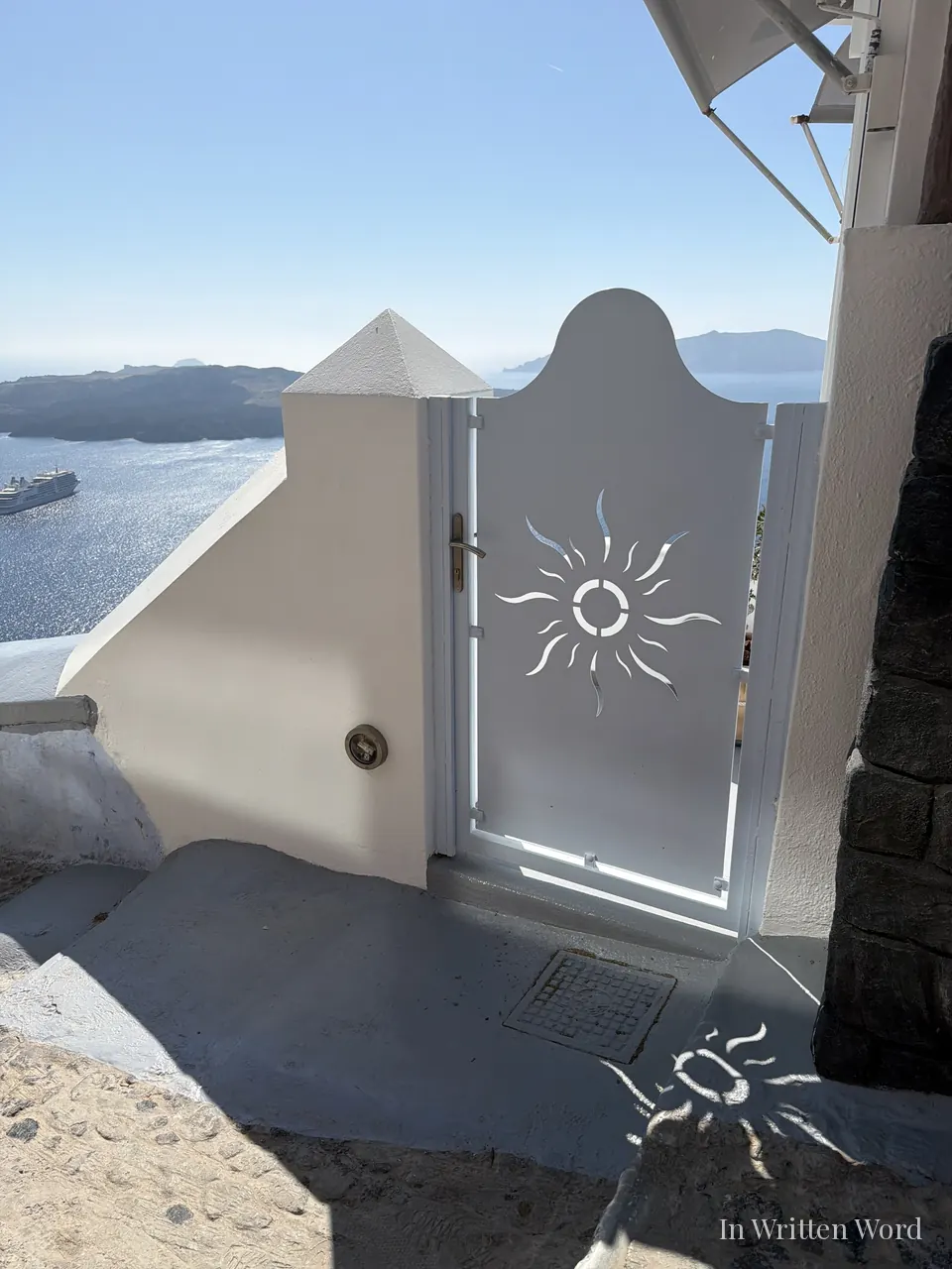 A white metal gate with a sun cutout design opening onto the Santorini caldera, with the sea, the distant volcanic islands, and a cruise ship visible in the bright haze beyond.