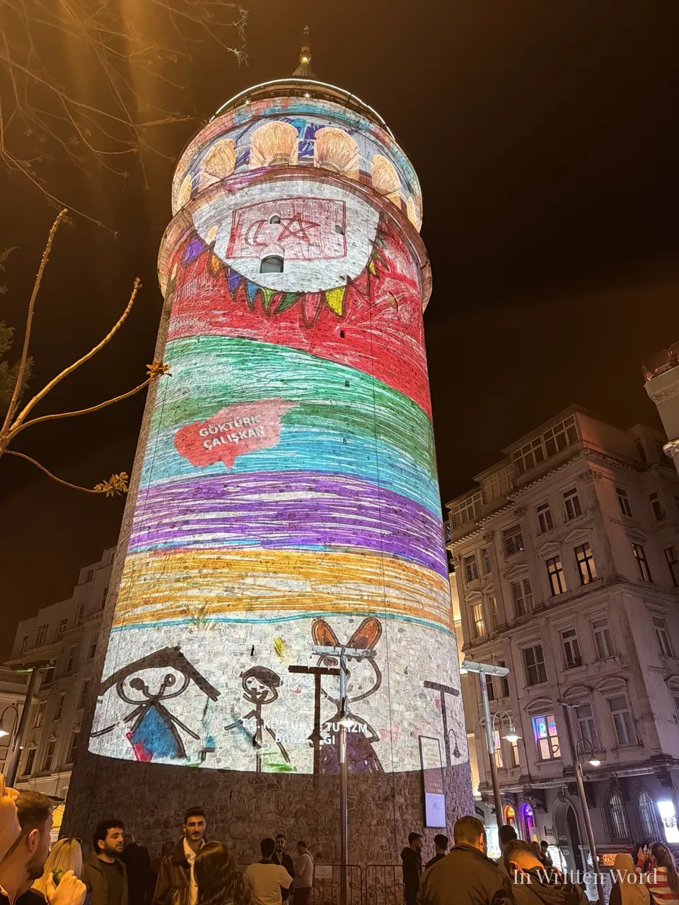 The medieval Galata Tower in Istanbul illuminated at night with a colorful projection of children’s crayon drawings across its stone facade, a crowd gathered below