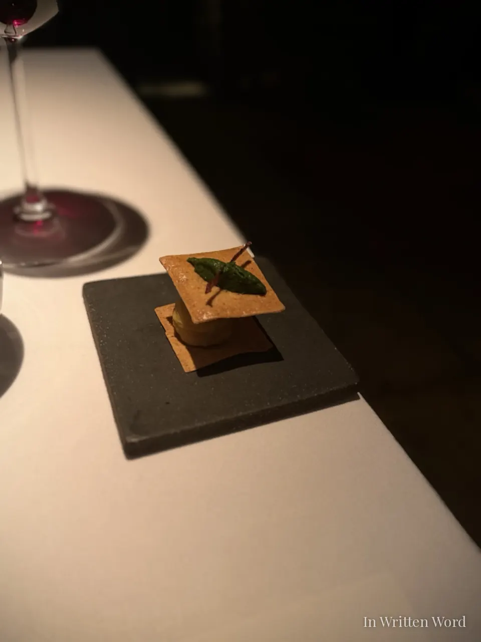 A delicate amuse-bouche served on a dark grey slate plate at Aheste restaurant in Istanbul, a single herb leaf placed precisely on top, with a wine glass visible in the background