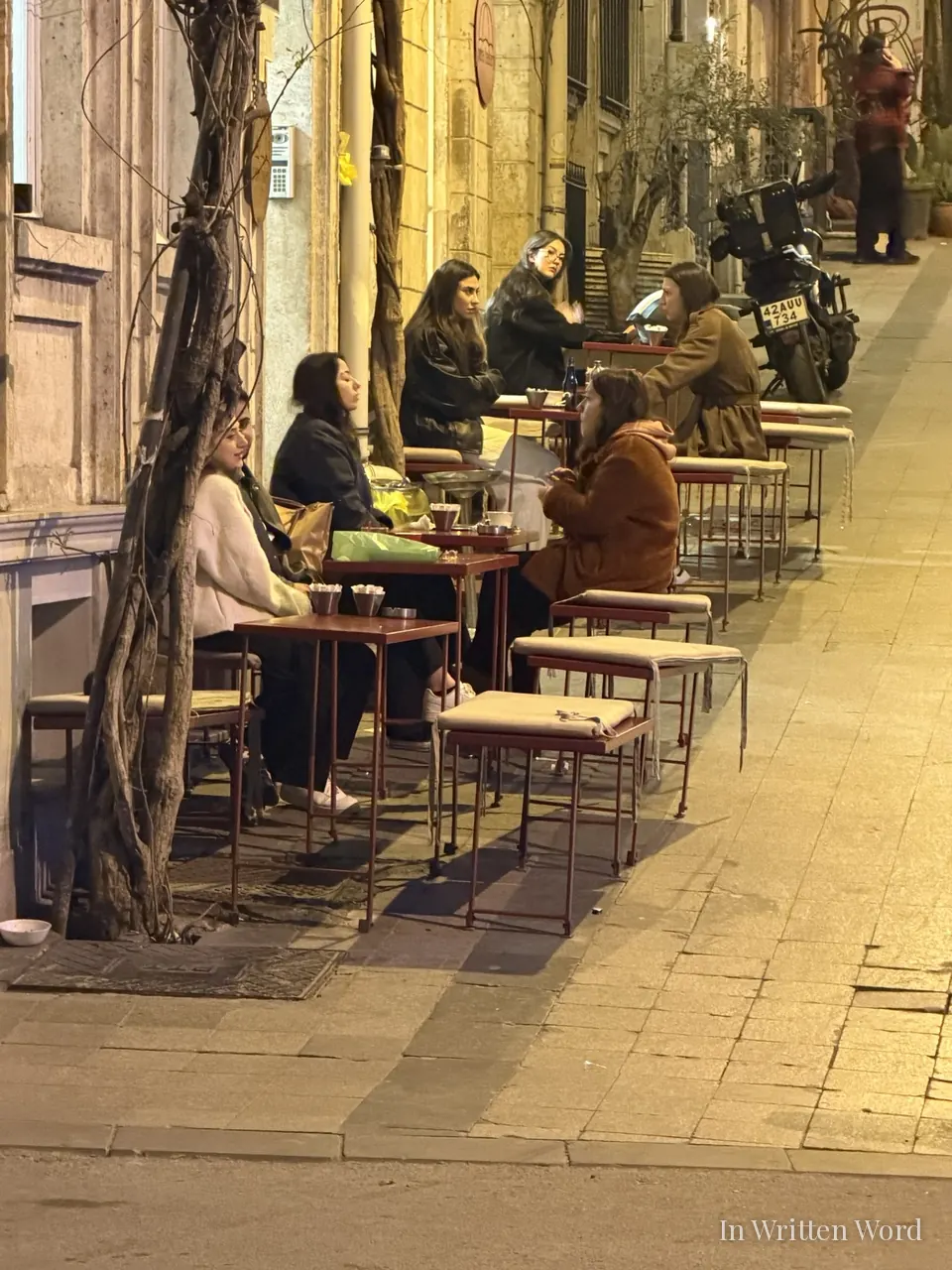 Women sitting at outdoor café tables on a quiet cobblestone street in Pera, Istanbul, at night, in warm amber light