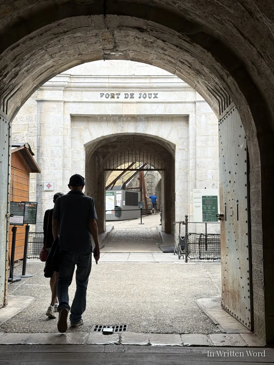 Two visitors walk through a stone archway into Fort de Joux, the fort's name carved in the pale stone above the inner gate.