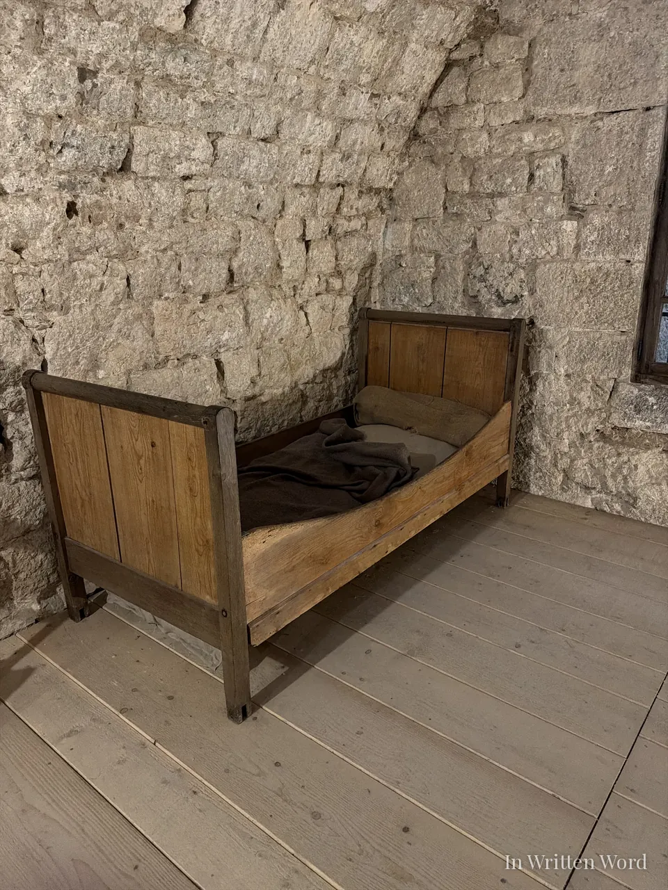 A bare wooden bed with a rough dark blanket in a cold stone cell at Fort de Joux, worn plank floor beneath it.