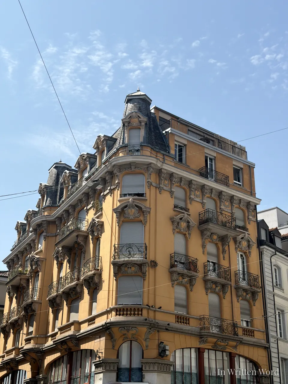An ornate mustard-yellow Belle Epoque corner building with wrought-iron balconies and a turret, against a clear blue sky in Lausanne.