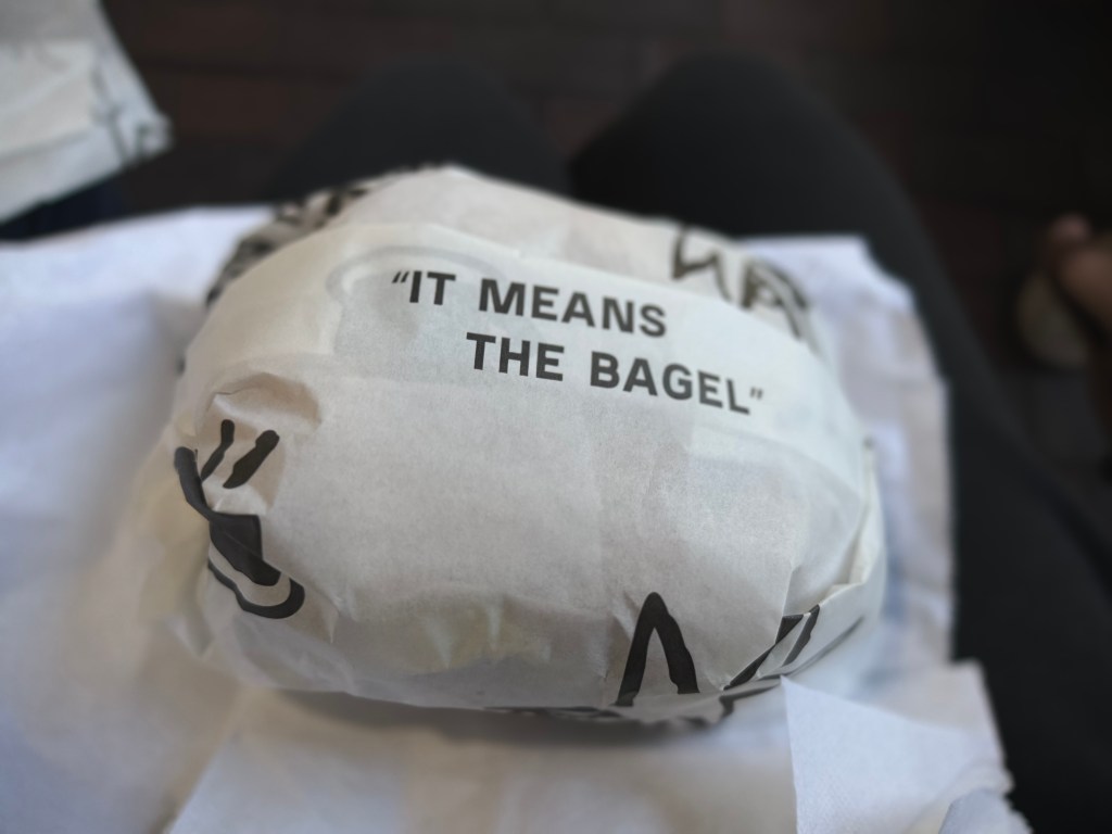 Bagel sandwich wrapped in white paper printed with the phrase “It means the bagel,” resting on a napkin.