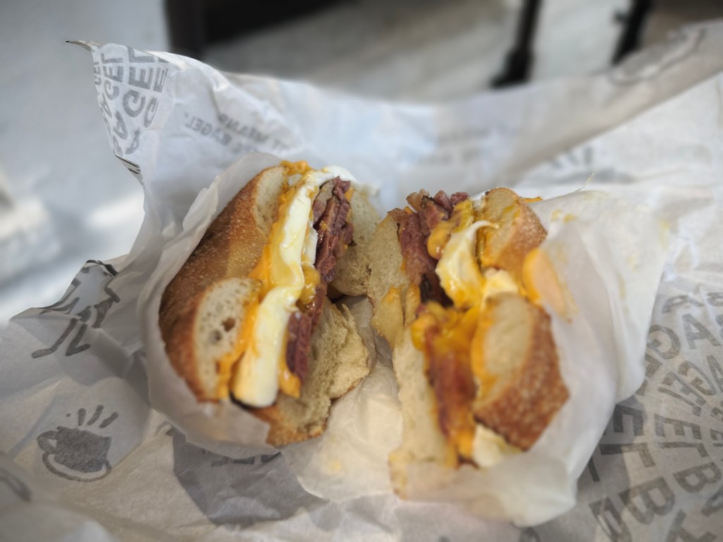 Close-up of a breakfast bagel sandwich cut in half, filled with egg, melted cheese, and meat, wrapped in paper.