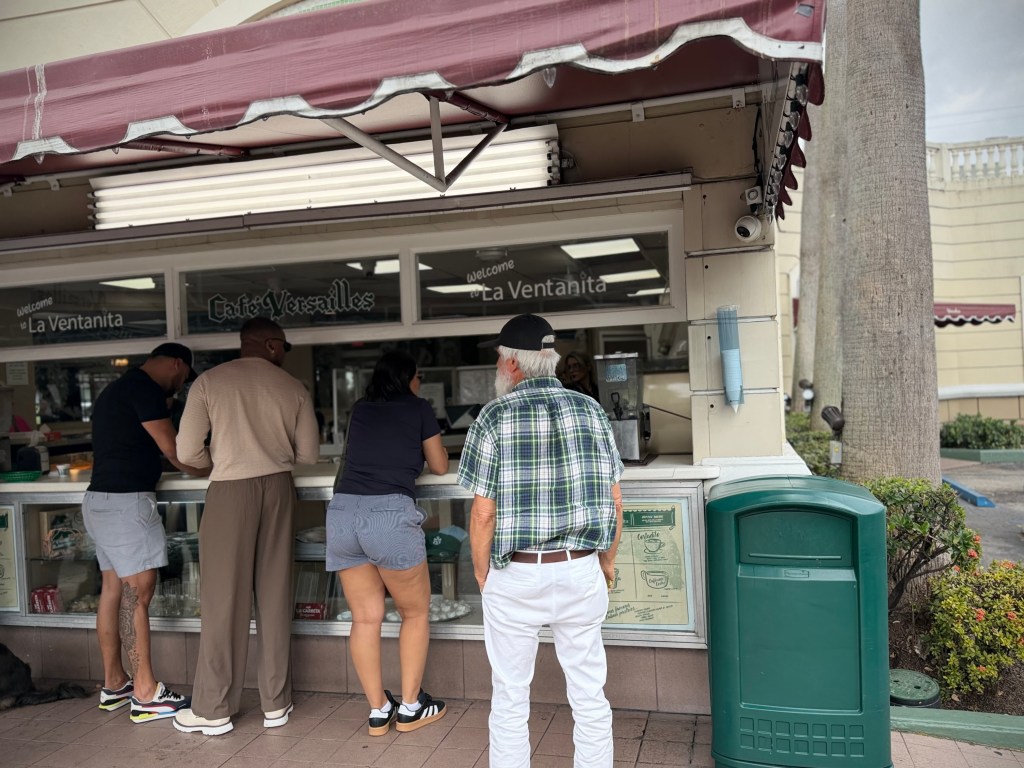 Customers standing at the outdoor coffee window at Versailles Restaurant in Miami, ordering at La Ventanita beneath a maroon awning.